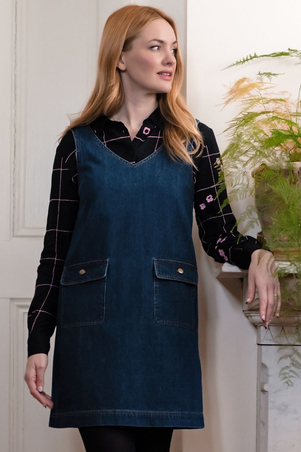 A woman wearing a dark blue denim apron over a black plaid shirt, standing indoors next to a potted plant.