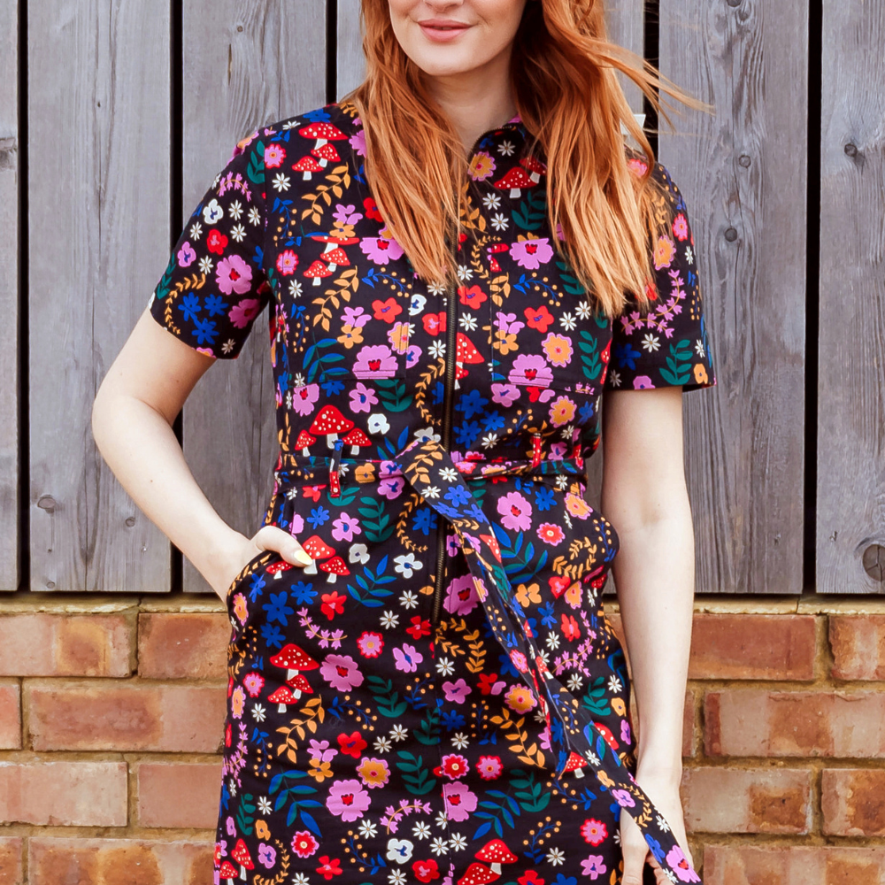 Woman wearing a colorful floral dress standing against a wooden and brick wall.