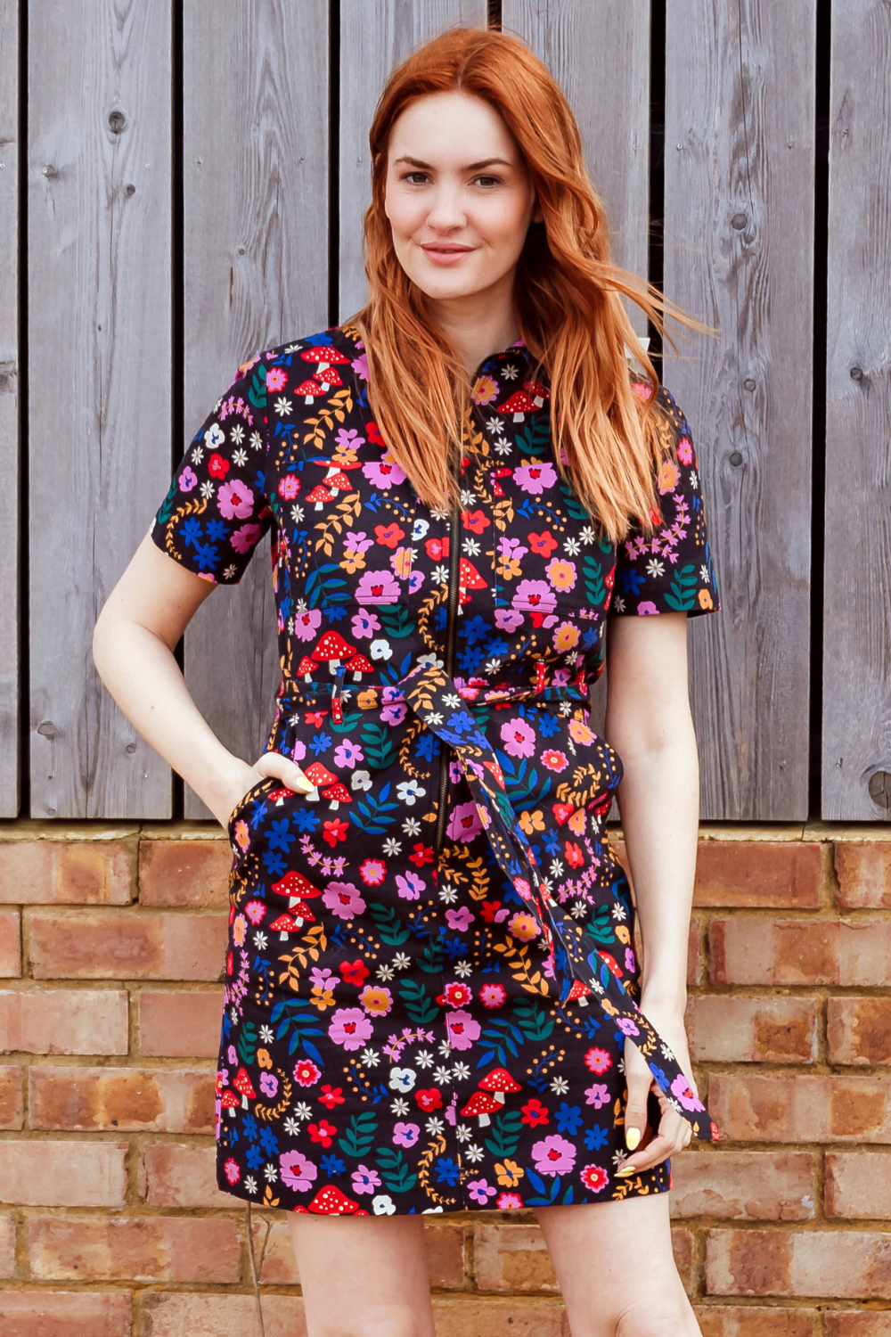 Woman wearing a colorful floral dress standing against a wooden and brick wall.