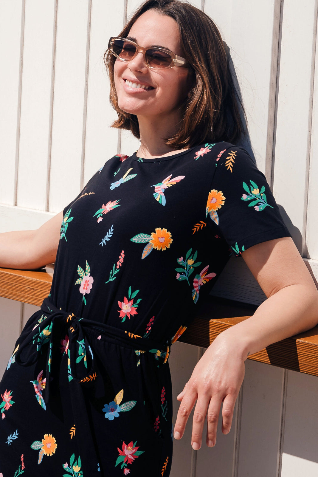 A woman smiling and wearing a black mini dress with a colorful hummingbird and flower design, featuring short sleeves and an elasticated waist.