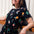 A woman smiling and wearing a black mini dress with a colorful hummingbird and flower design, featuring short sleeves and an elasticated waist.