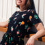 A woman smiling and wearing a black mini dress with a colorful hummingbird and flower design, featuring short sleeves and an elasticated waist.