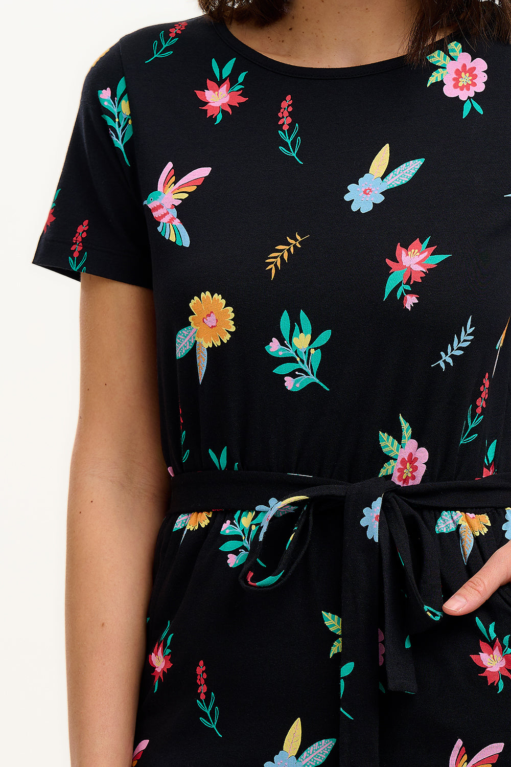 A model wearing a short-sleeved black mini dress with a multicolored hummingbird and garden design, standing against a white background.