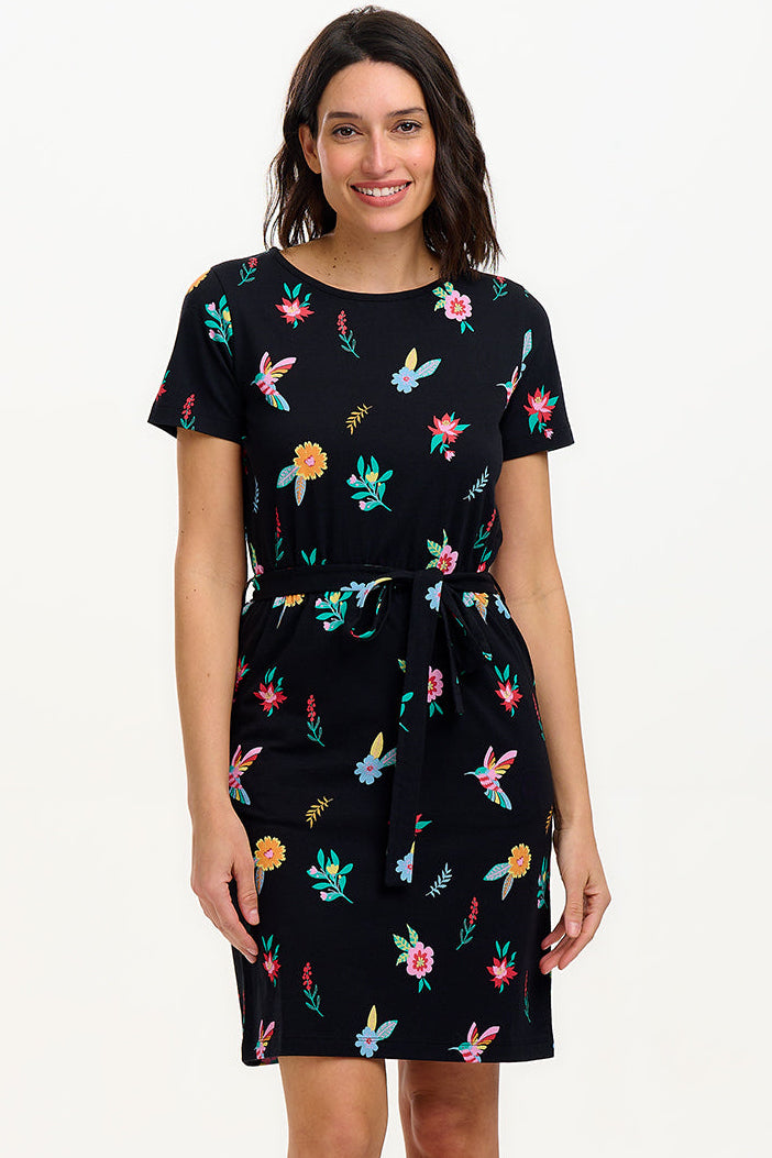 A model wearing a short-sleeved black mini dress with a multicolored hummingbird and garden design, standing against a white background.