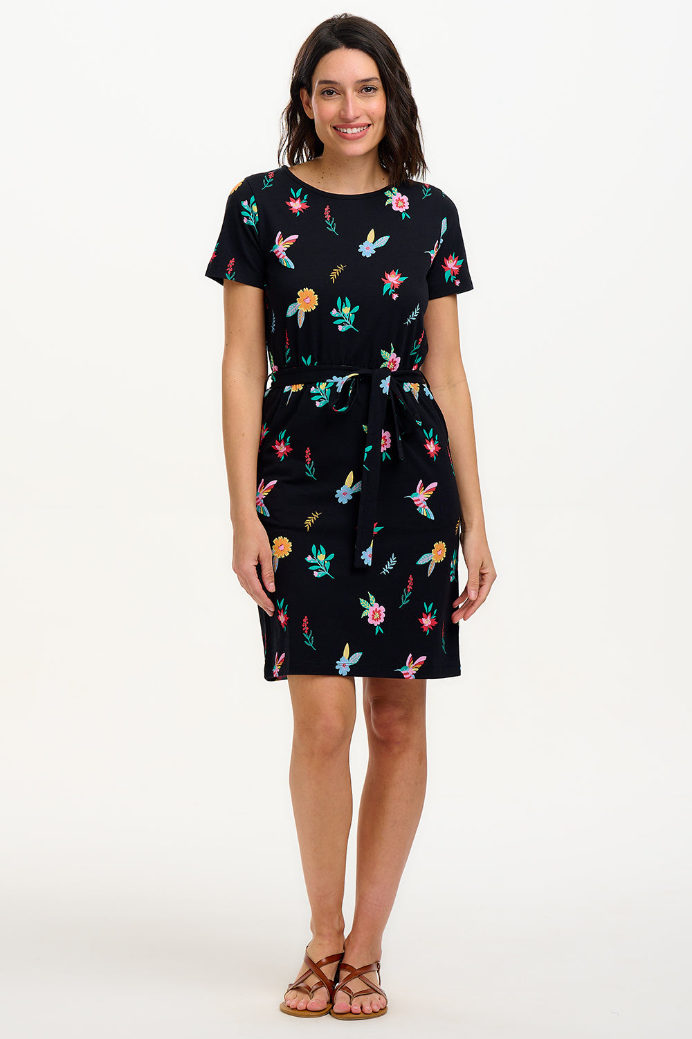 A model wearing a short-sleeved black mini dress with a multicolored hummingbird and garden design, standing against a white background.