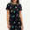 A model wearing a short-sleeved black mini dress with a multicolored hummingbird and garden design, standing against a white background.