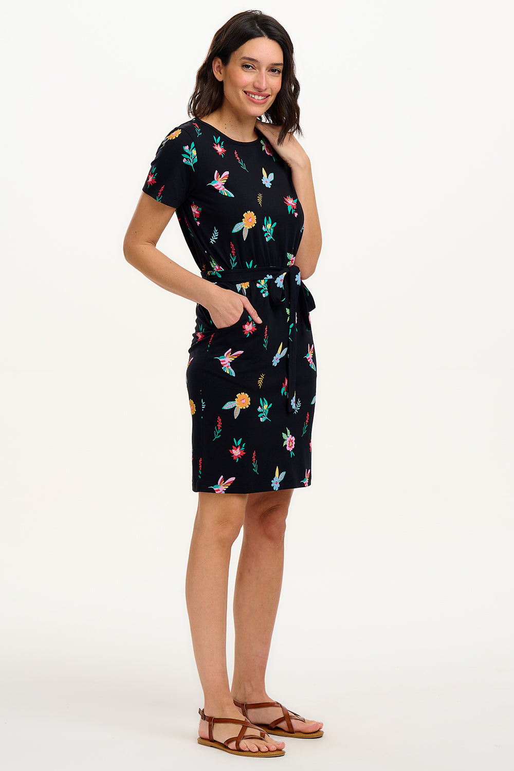 A model wearing a short-sleeved black mini dress with a multicolored hummingbird and garden design, standing against a white background.