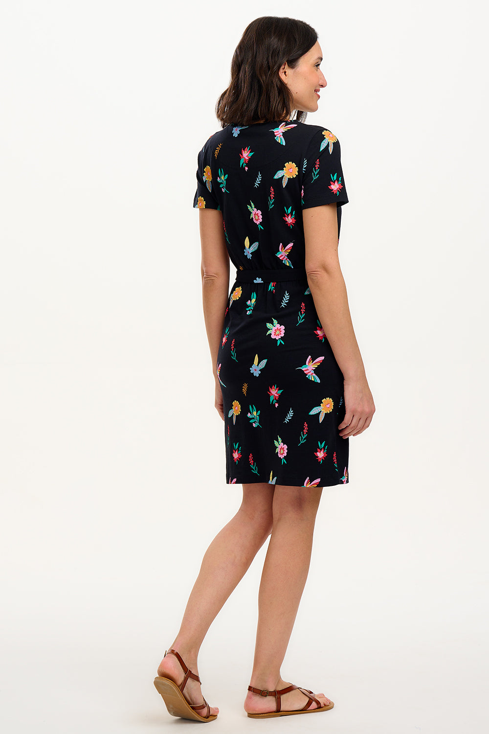 A model wearing a short-sleeved black mini dress with a multicolored hummingbird and garden design, standing against a white background.