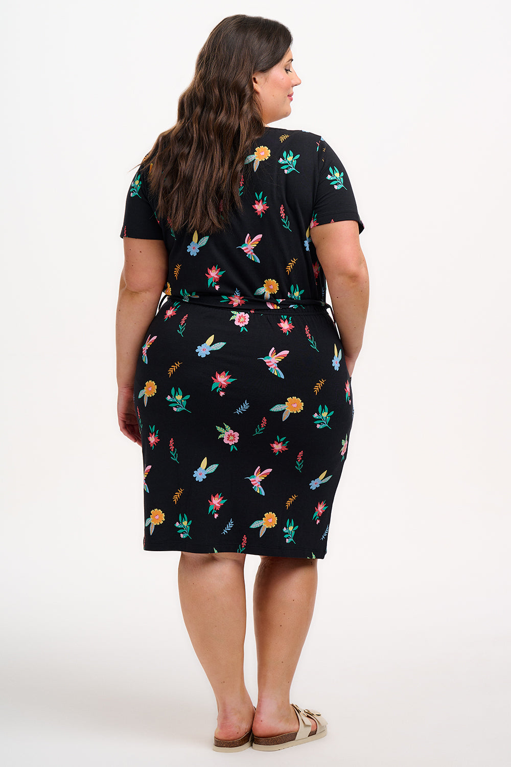 A model wearing a short-sleeved black mini dress with a multicolored hummingbird and garden design, standing against a white background.