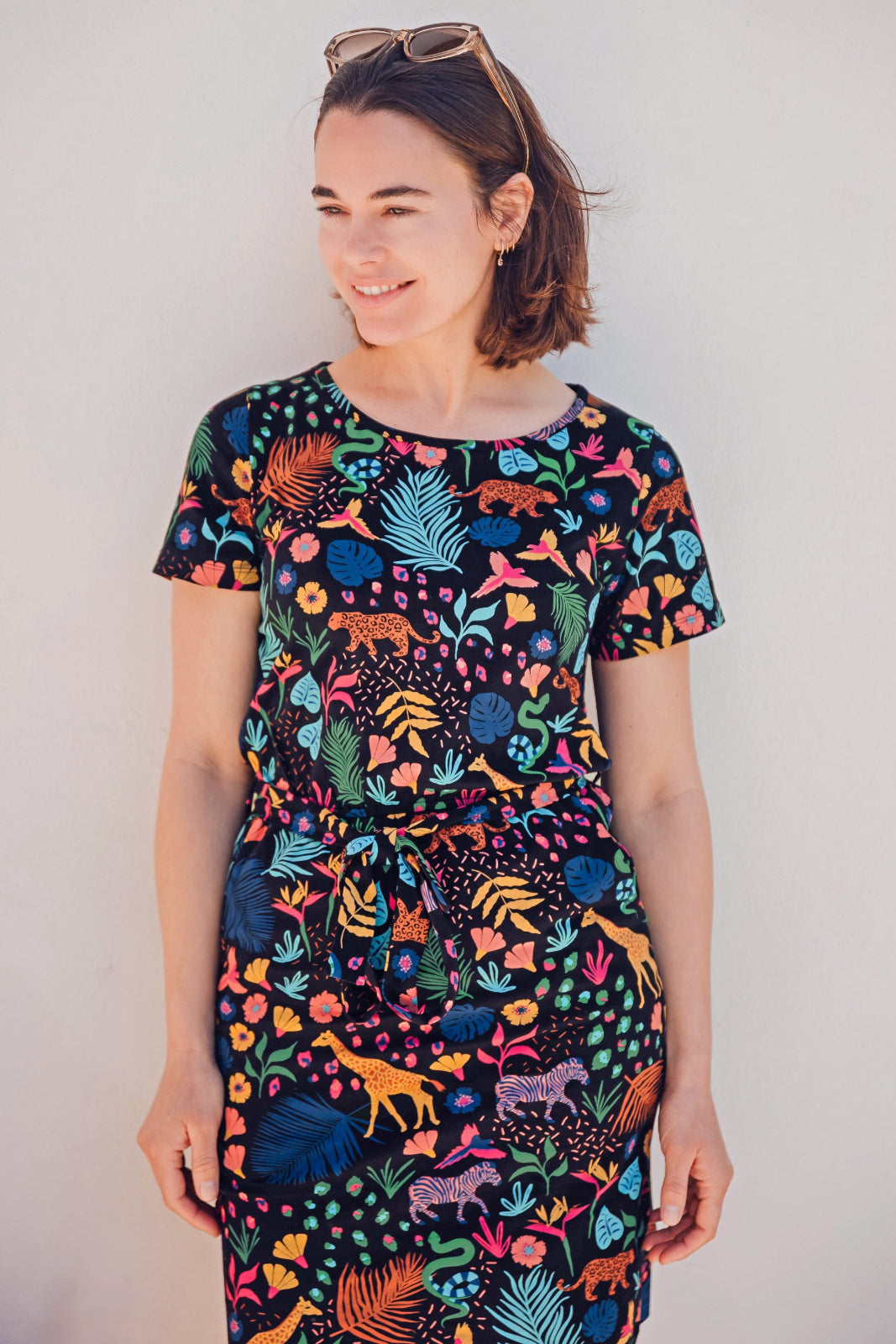 A model wearing a short-sleeved mini dress with a colorful jungle animal and foliage pattern on a black background.