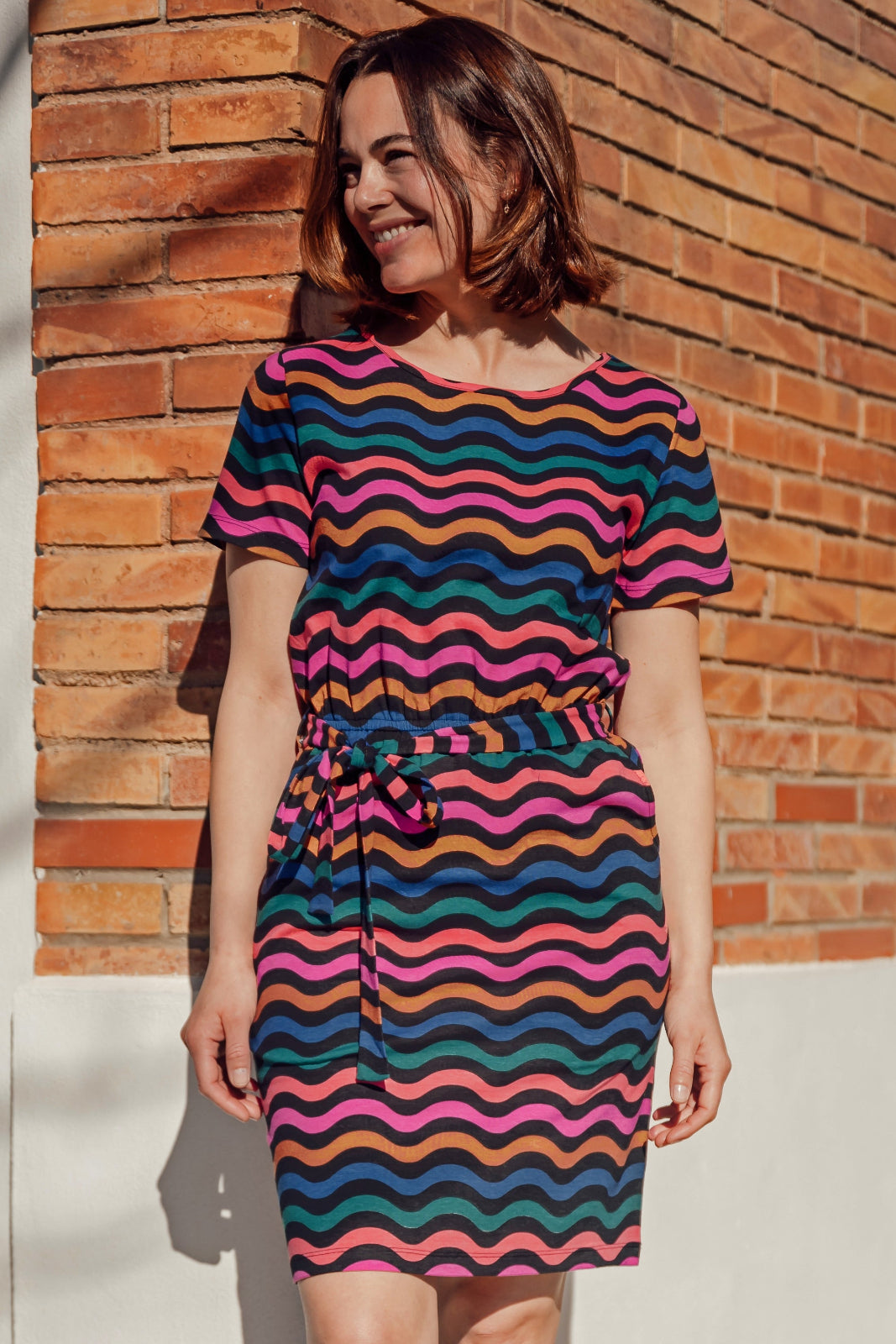 A model wearing a short-sleeved mini dress with wavy chevron stripes in black, teal, navy, tan, pink, and coral colors, standing against a brick wall background.
