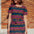A model wearing a short-sleeved mini dress with wavy chevron stripes in black, teal, navy, tan, pink, and coral colors, standing against a brick wall background.