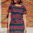 A model wearing a short-sleeved mini dress with wavy chevron stripes in black, teal, navy, tan, pink, and coral colors, standing against a brick wall background.