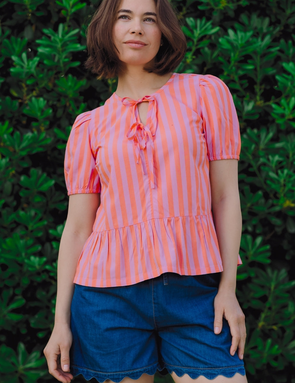 Woman wearing a pink and white striped top with blue shorts against a green leafy background