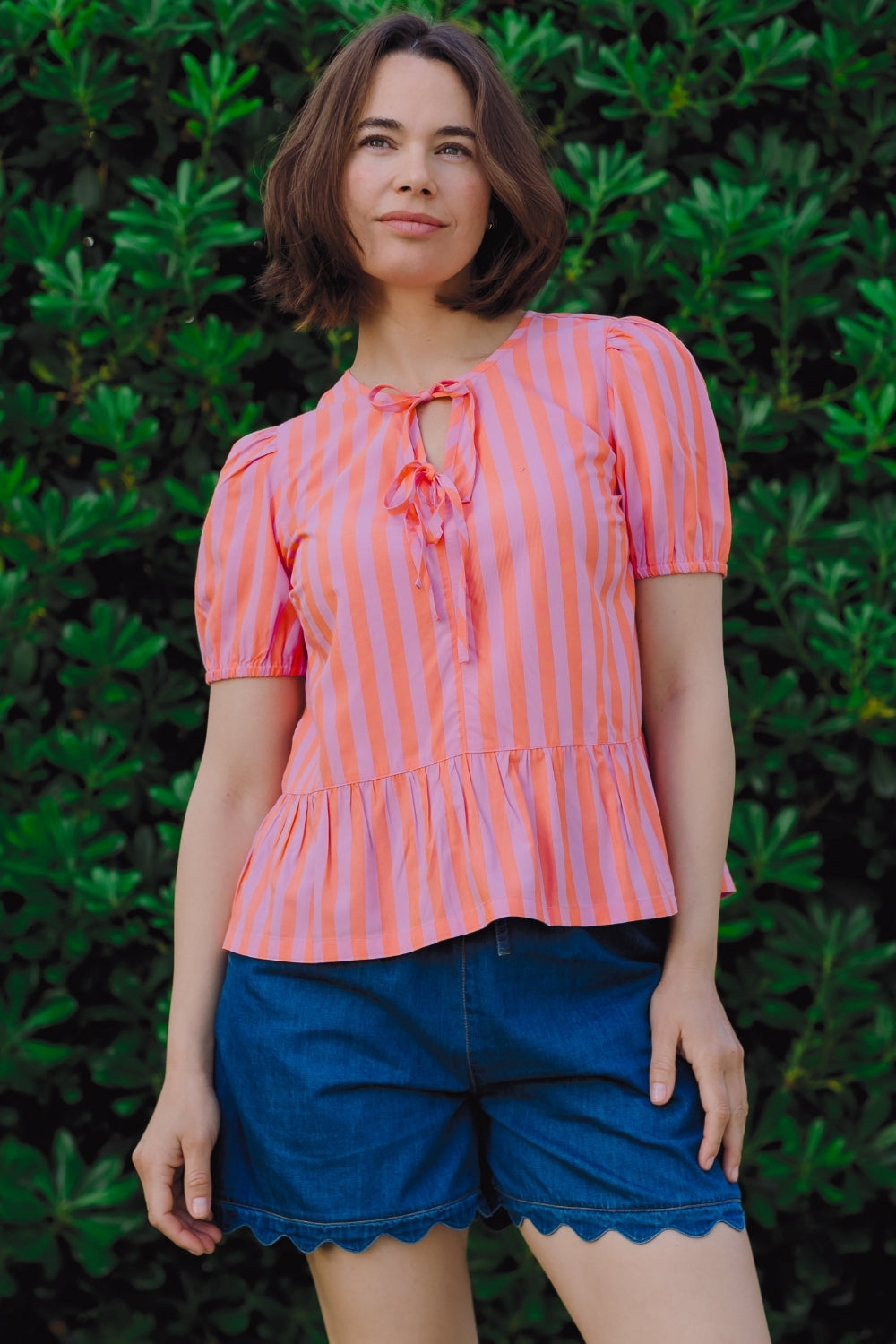 Woman wearing a pink and white striped top with blue shorts against a green leafy background