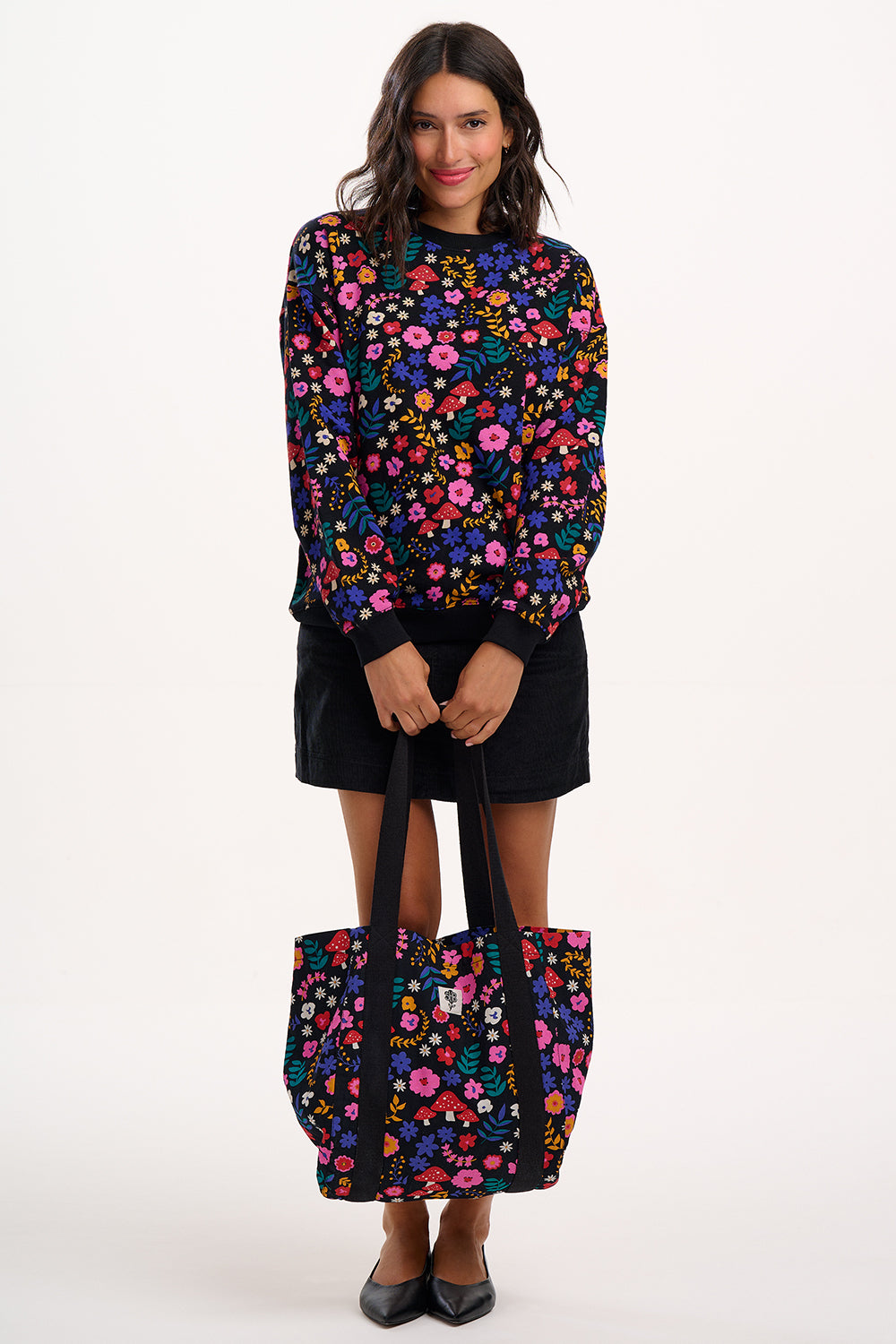 Woman wearing a colorful floral sweater holding a matching tote bag on a white background