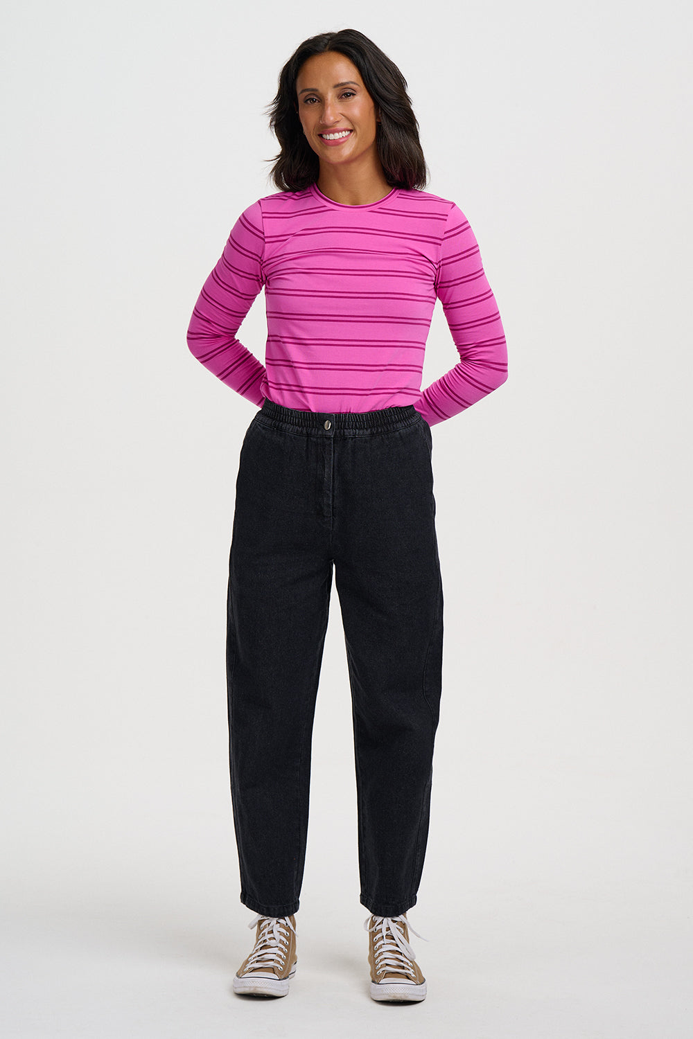 A woman modeling a bright pink long-sleeve striped t-shirt paired with black jeans and beige sneakers, standing against a plain white studio background.