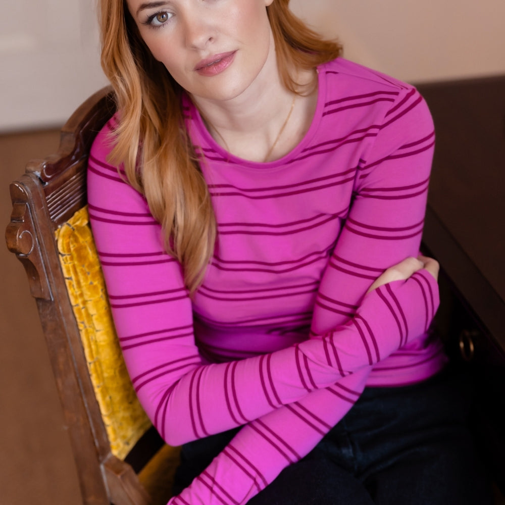 A woman with red hair sits on a yellow upholstered wooden chair, wearing a bright pink long-sleeve top with thin dark horizontal stripes and dark pants.
