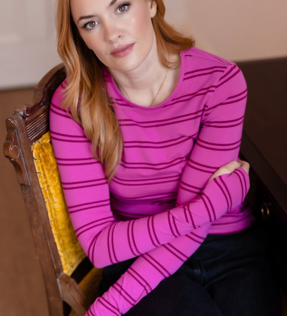 A woman with red hair sits on a yellow upholstered wooden chair, wearing a bright pink long-sleeve top with thin dark horizontal stripes and dark pants.