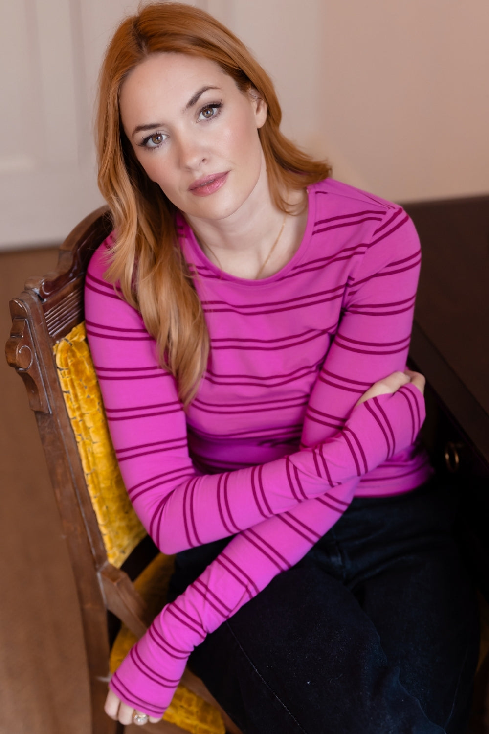 A woman with red hair sits on a yellow upholstered wooden chair, wearing a bright pink long-sleeve top with thin dark horizontal stripes and dark pants.