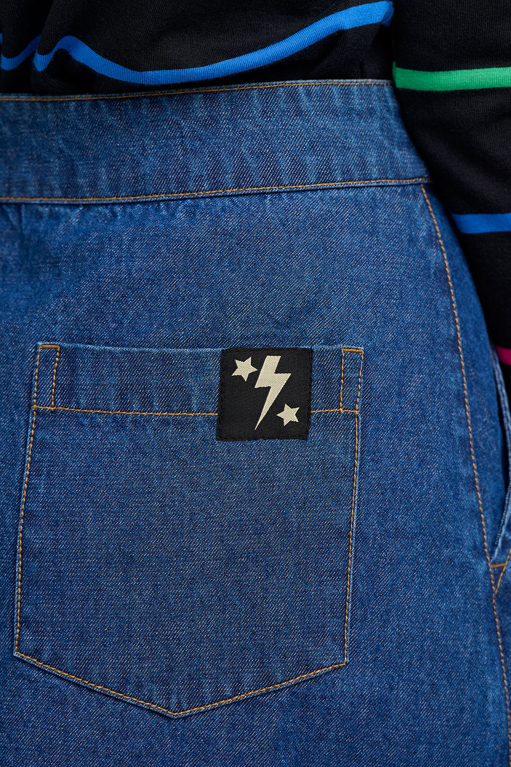 Suagrhill Brighton blue denim apron with a black patch featuring stars and a lightning bolt on a person wearing a black and green jacket.
