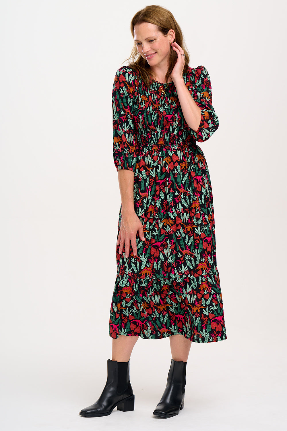 A woman standing and posing in a floral midi dress with an elasticated shirred bust and full midi skirt, featuring a hand-drawn 'Lost Dinosaur' print.