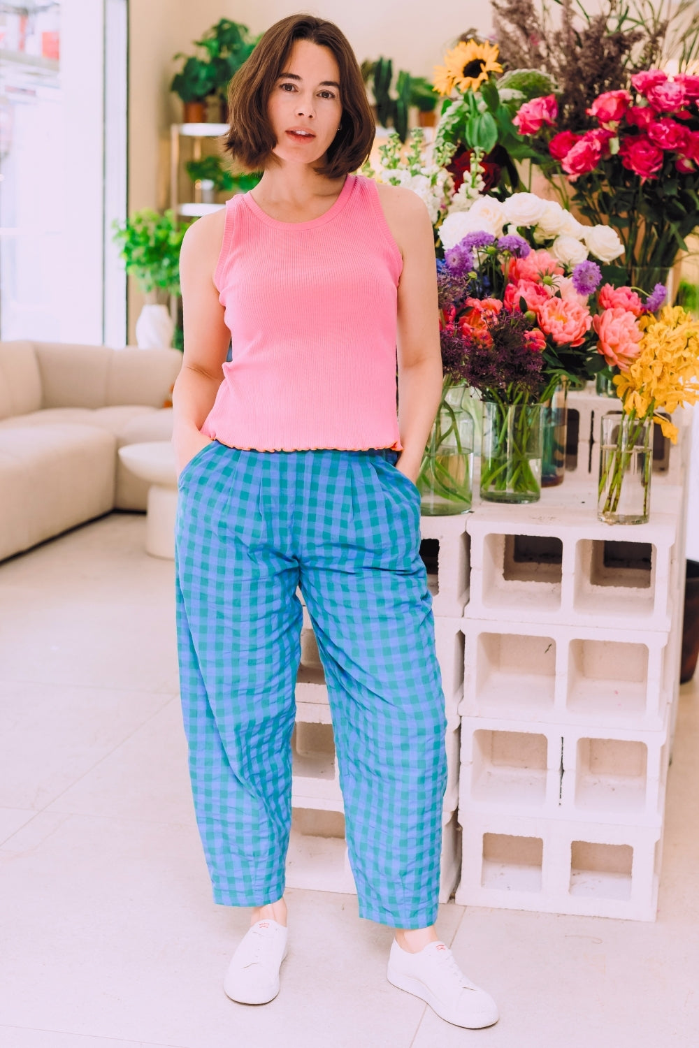 Woman in pink top and blue checkered pants standing in a room with flowers and a white couch.