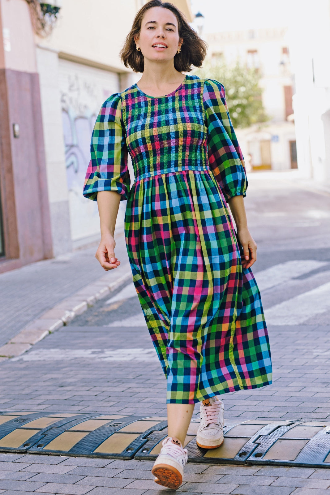 A woman walking on a street wearing a midi dress with a shirred bodice and 3/4 length sleeves in a checkered pattern.
