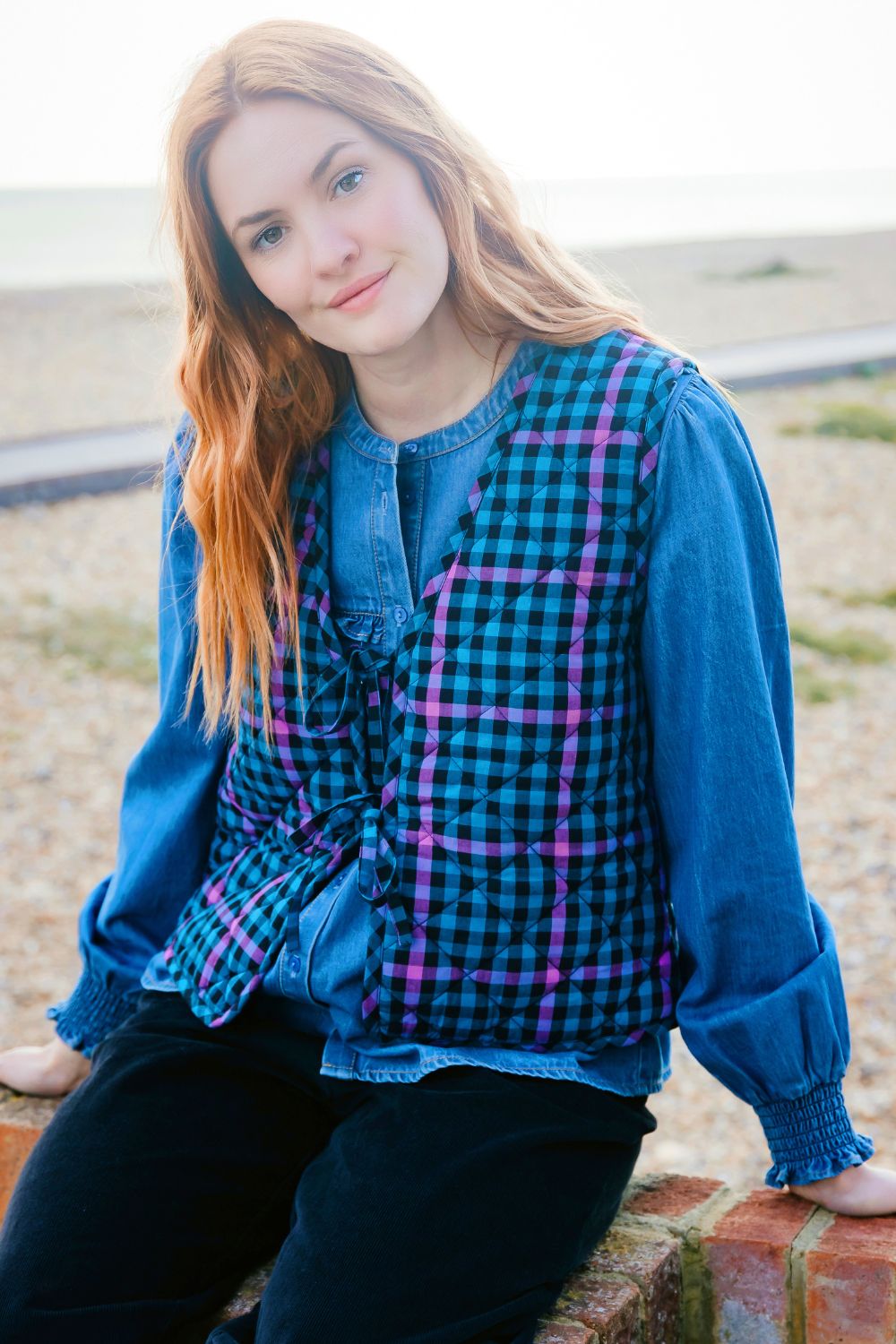 Woman wearing a blue plaid waistcoat with pink accents sitting on a brick wall.