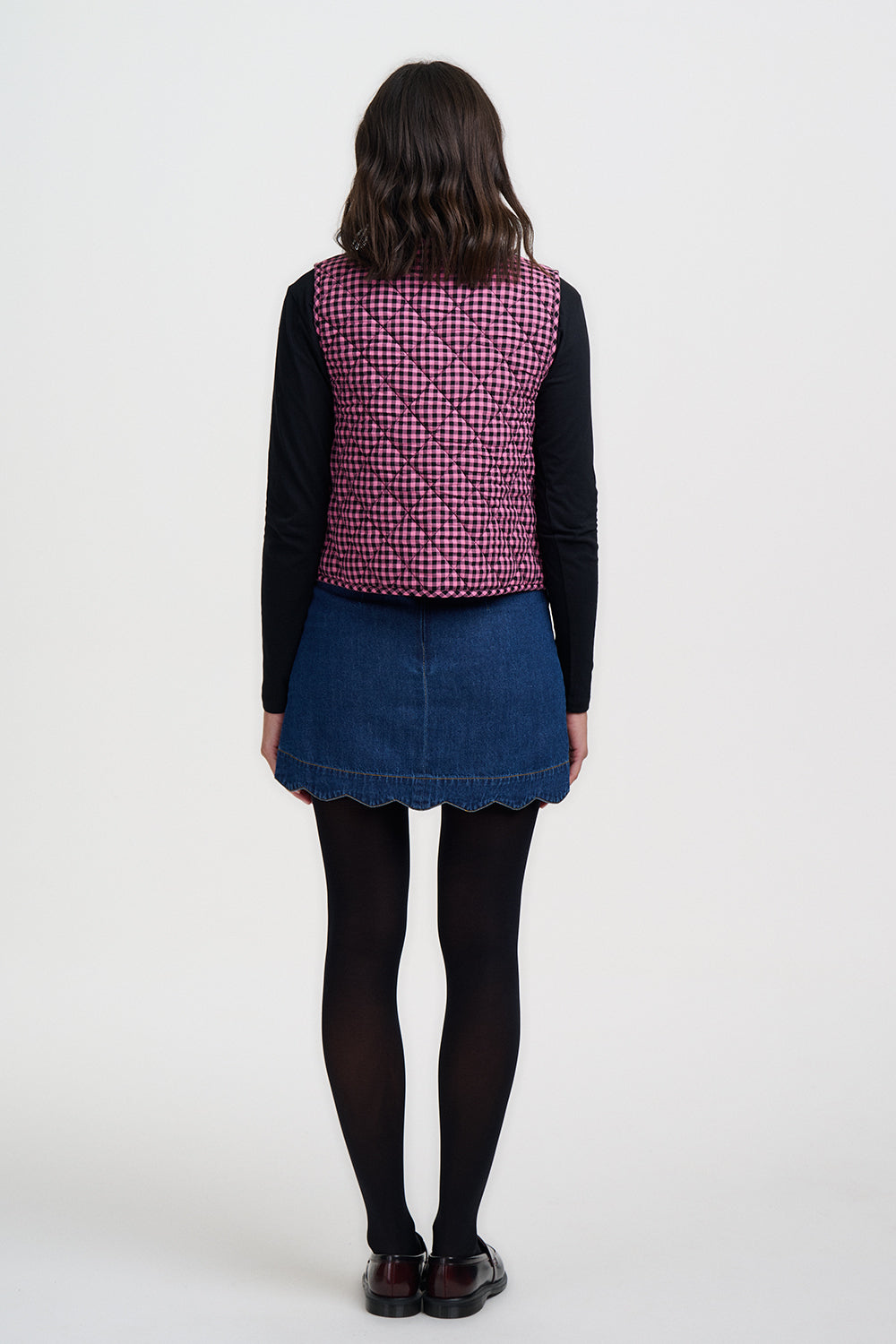 Person wearing a pink checkered vest over a black long-sleeve shirt and blue denim skirt on a white background