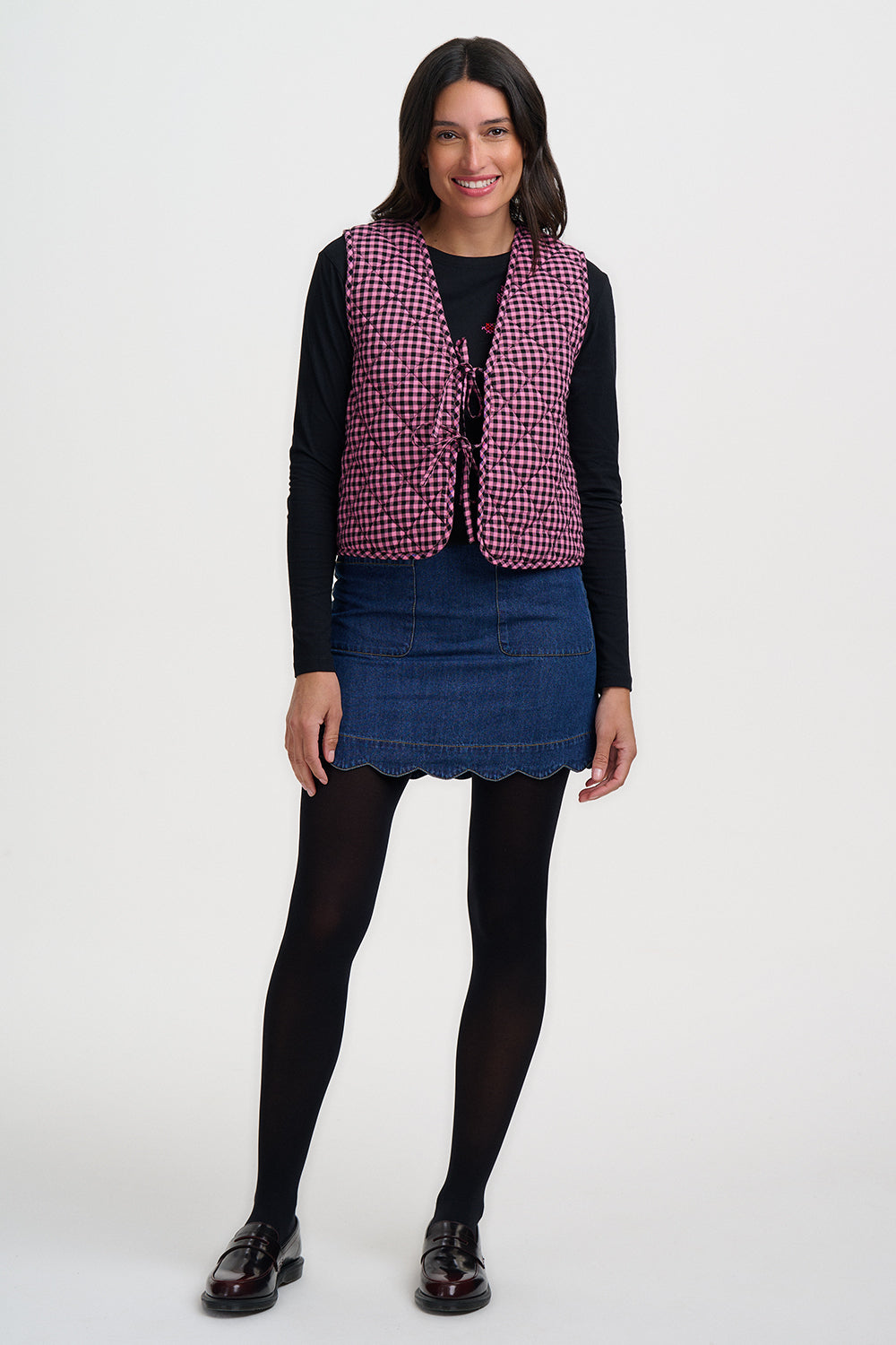 Woman wearing a pink checkered vest over a black long-sleeve shirt and blue skirt on a white background