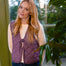 A woman with long blonde hair wearing a purple and white gingham tie-front vest over a cream long-sleeve top and dark blue jeans, standing indoors near a window with green foliage in the background.