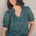 A woman smiling and wearing a green blouse with a geometric concentric circles pattern, featuring a frill collar and v-neckline with ties, short puff sleeves with elasticated frill cuffs.