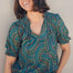A woman smiling and wearing a green blouse with a geometric concentric circles pattern, featuring a frill collar and v-neckline with ties, short puff sleeves with elasticated frill cuffs.