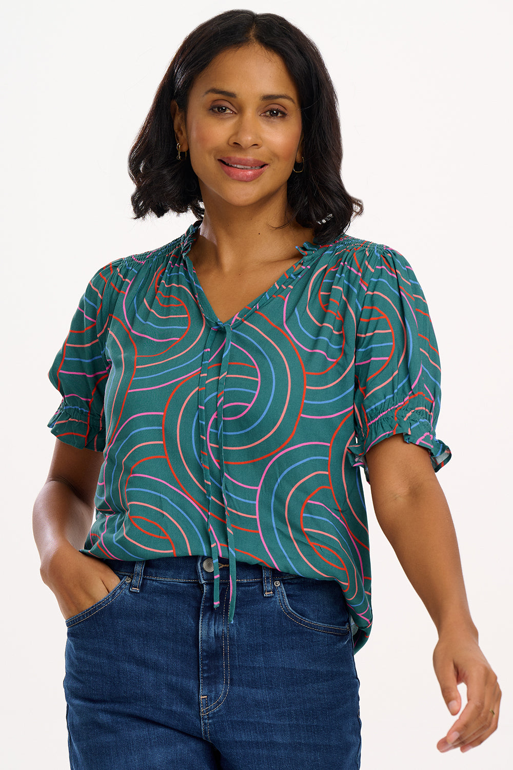 A woman modeling a green shirt with a geometric concentric circles pattern, featuring a frill collar and ties at the neckline, short puff sleeves with elasticated frill cuffs, and a v-neckline.