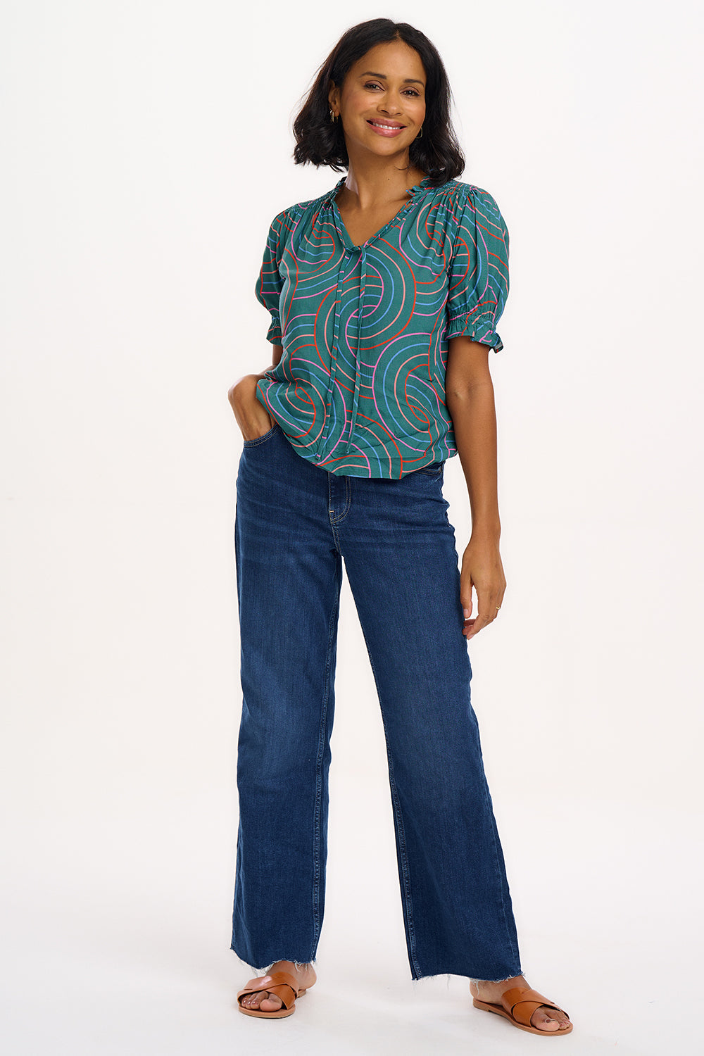 A woman smiling and wearing a green blouse with a geometric concentric circles pattern, featuring a frill collar and v-neckline with ties, short puff sleeves with elasticated frill cuffs.