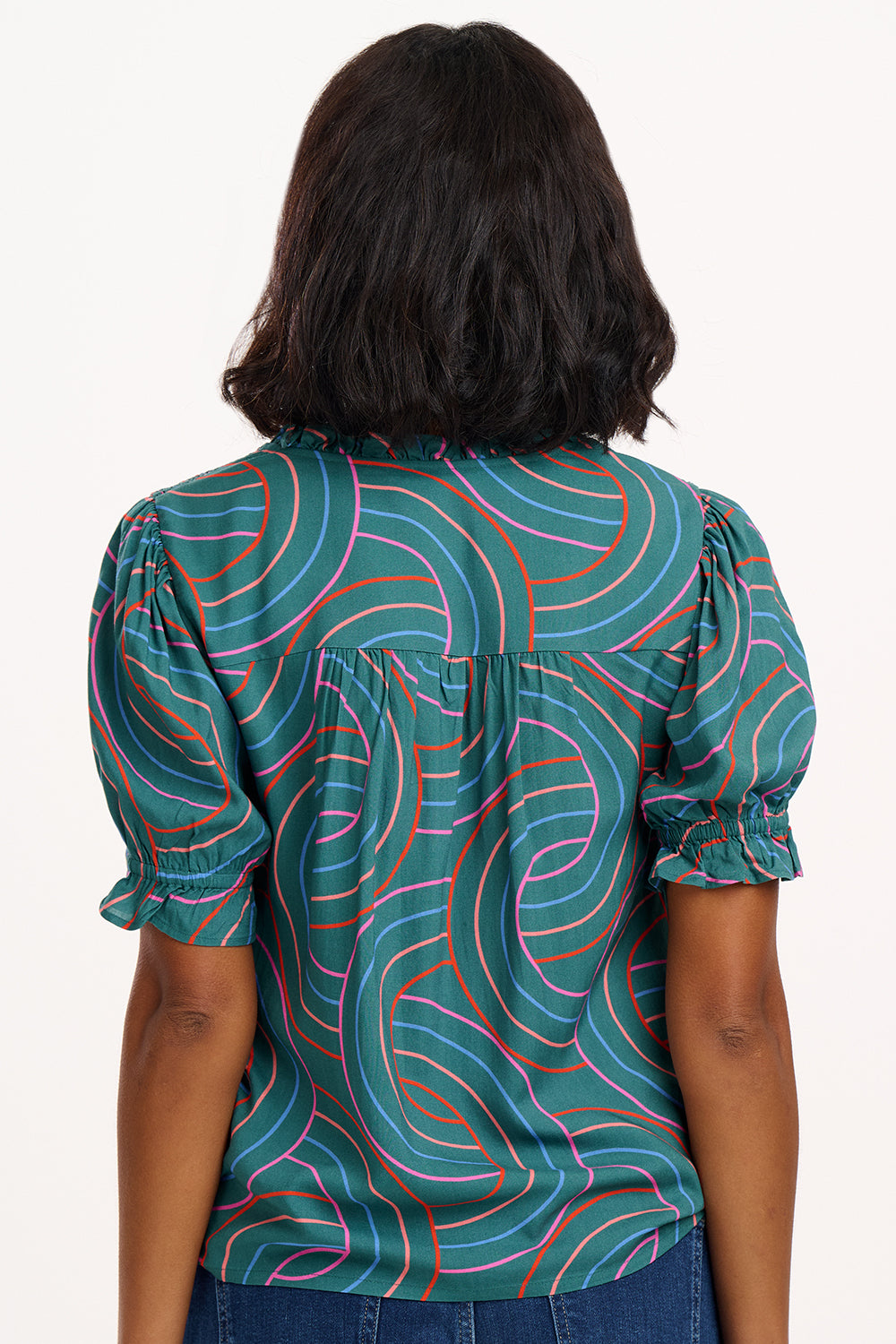 A woman smiling and wearing a green blouse with a geometric concentric circles pattern, featuring a frill collar and v-neckline with ties, short puff sleeves with elasticated frill cuffs.