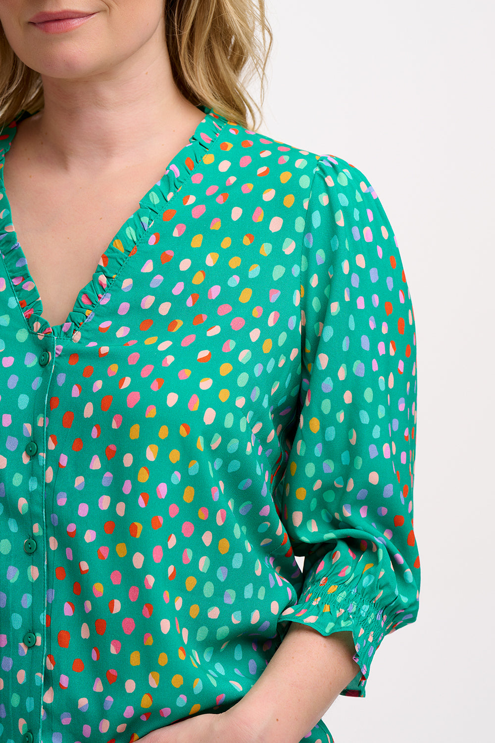 A woman wearing a green V-neck blouse with a pattern of rainbow pebbles, with long sleeves and a frill cuff.