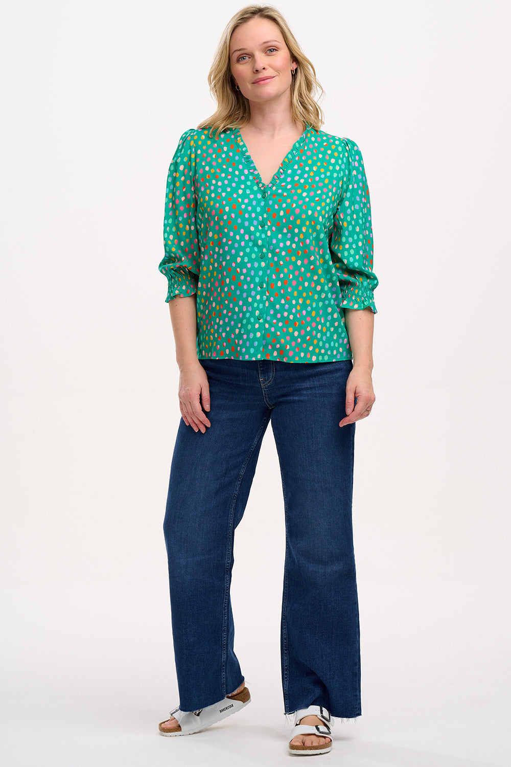 A woman wearing a green V-neck blouse with a pattern of rainbow pebbles, with long sleeves and a frill cuff.