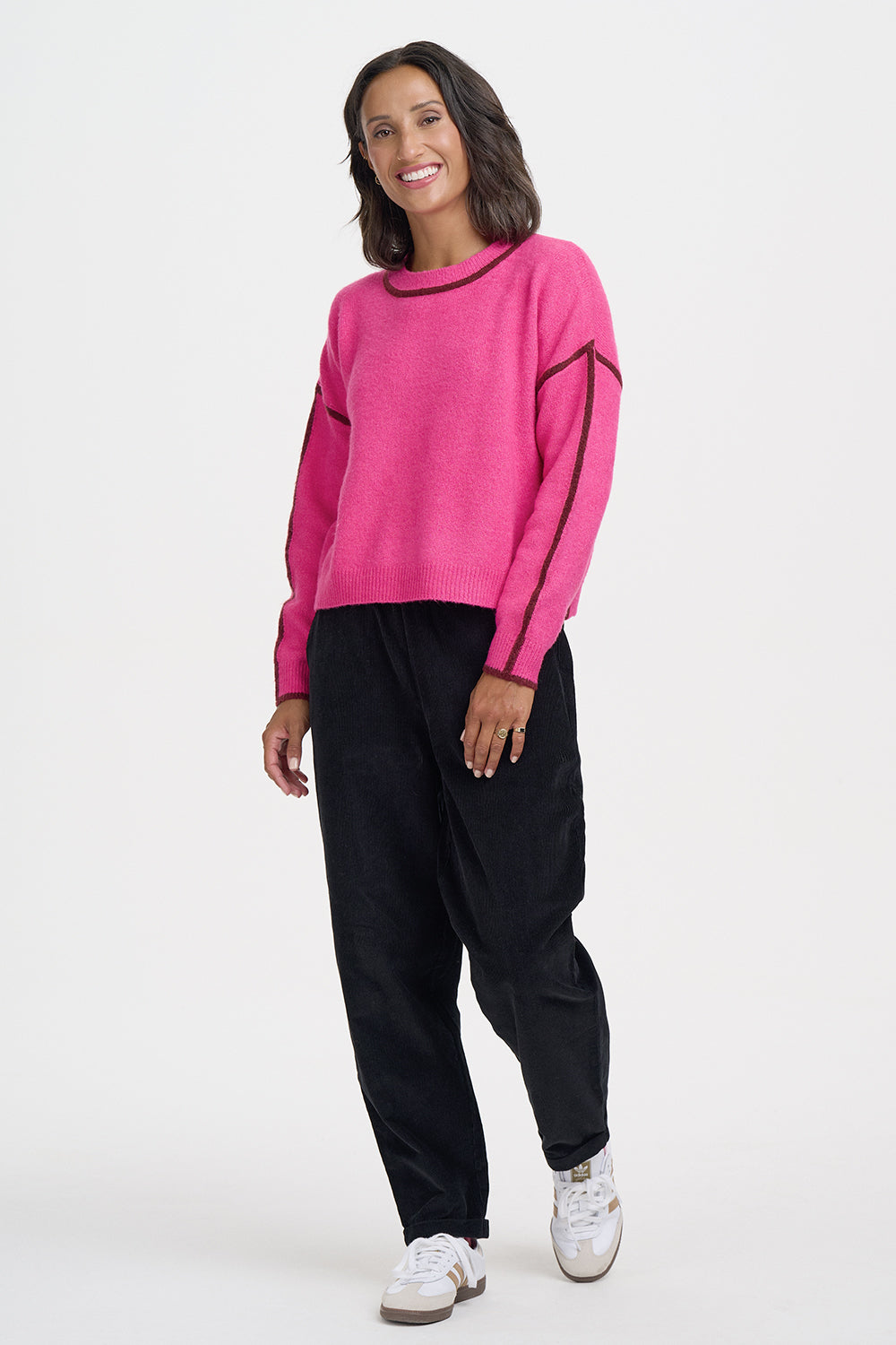 A woman modeling a bright pink knit sweater with black contrast trim and black jogger pants, standing against a plain light gray studio background.