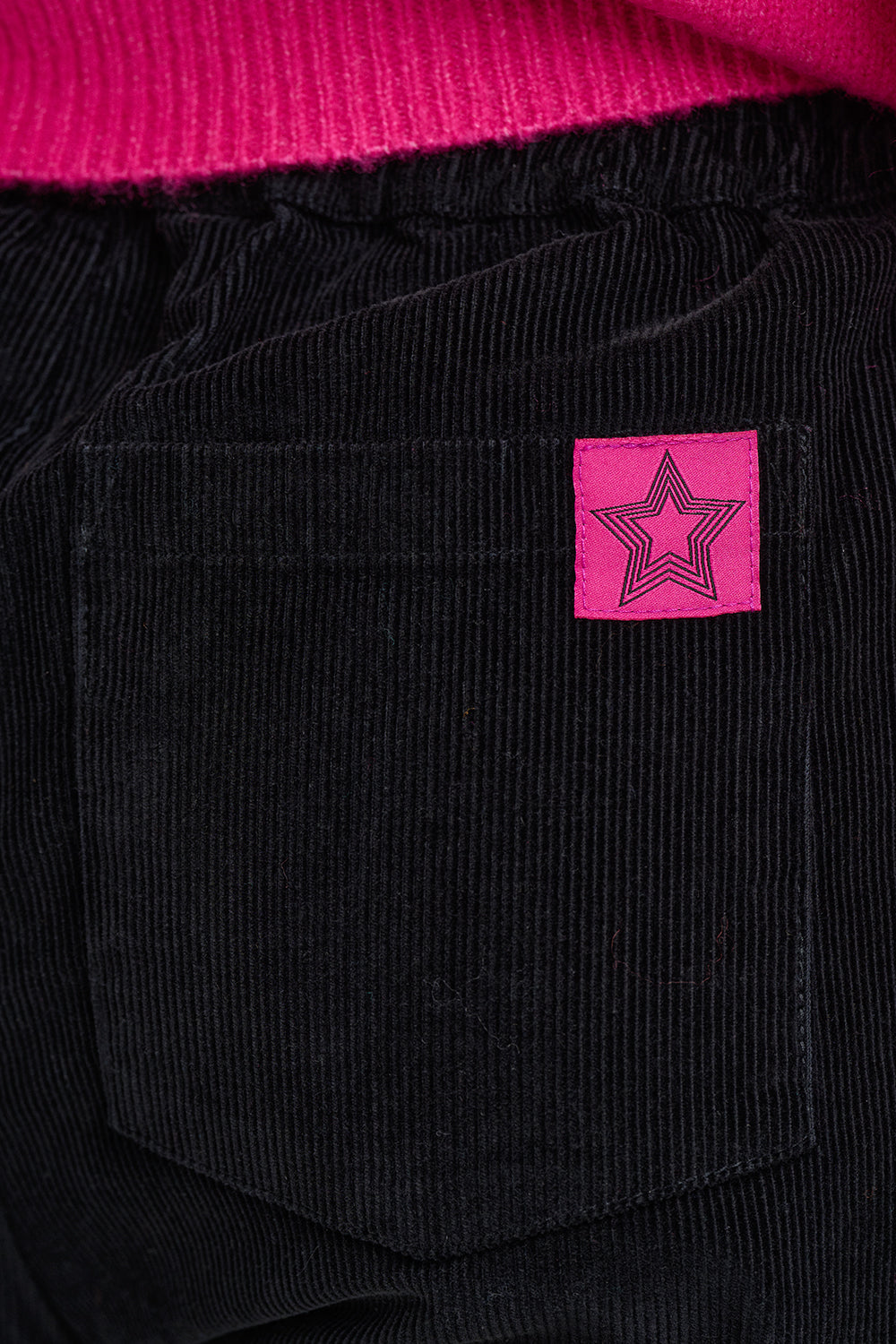 Close-up of black corduroy pants with a pink star patch on the back pocket, next to a bright pink ribbed knit garment.