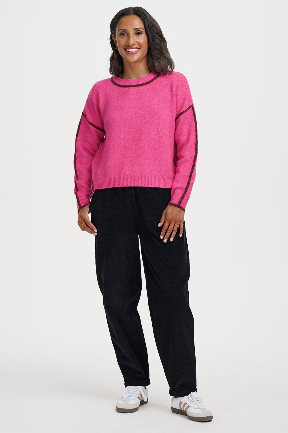A woman modeling a bright pink knit sweater with contrasting dark trim on the crew neck and sleeves, paired with black pants and white sneakers, against a plain light gray studio background.