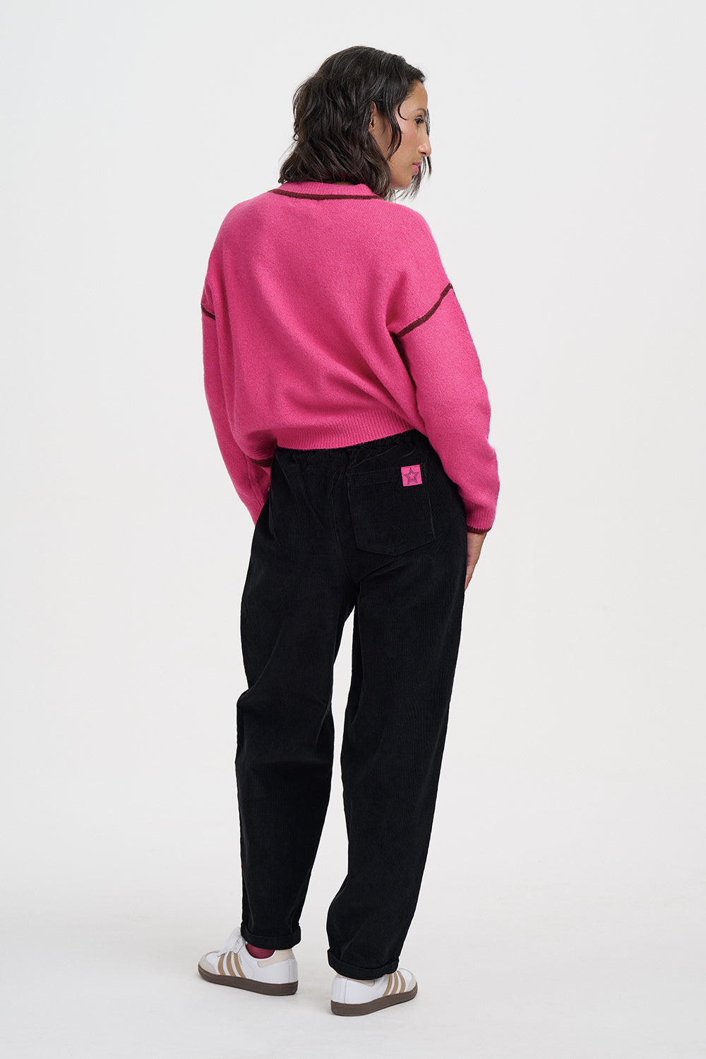 A model shown from the back wearing a bright pink cropped knit sweater with black contrast piping, black wide-leg pants, and white sneakers with pink stripes, against a plain white studio background.