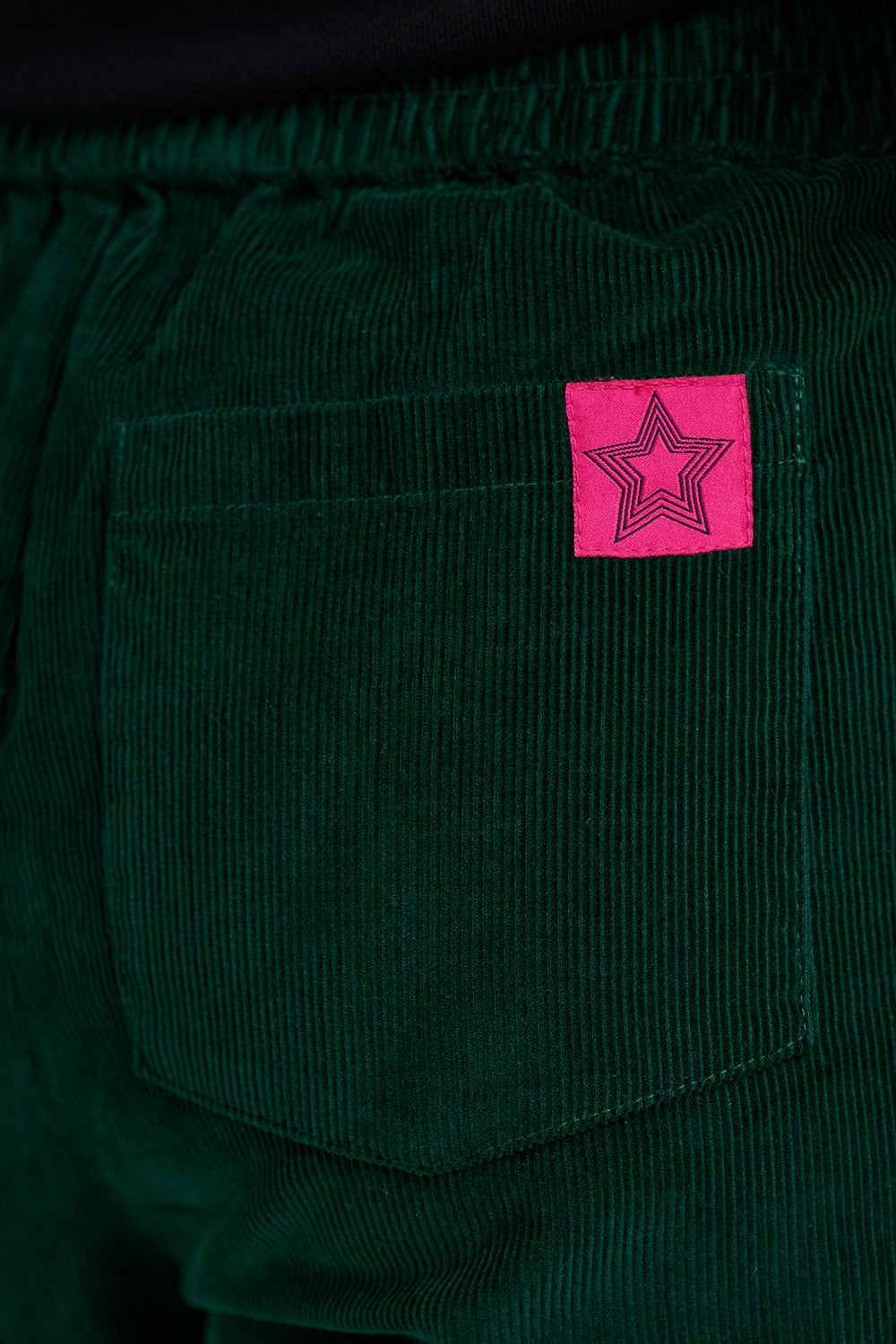 Close-up of dark green corduroy pants showing a pocket with a bright pink square patch featuring a black outlined star.