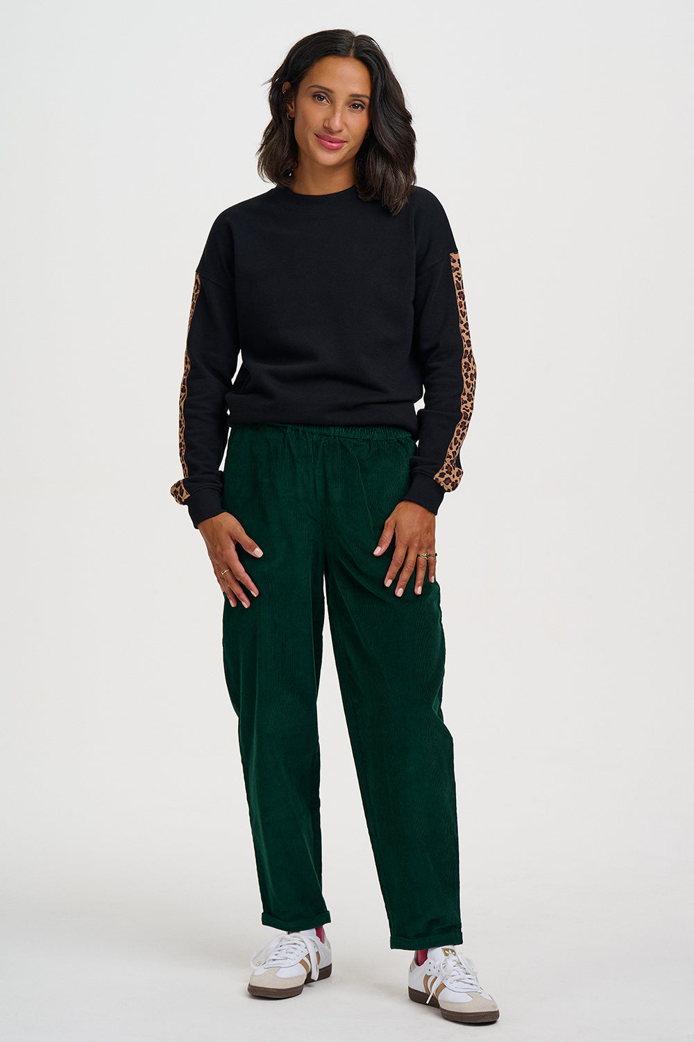 A woman modeling a black long-sleeve sweatshirt with leopard-print lace-up sleeves and dark green corduroy pants, paired with white sneakers, against a plain light gray studio background.