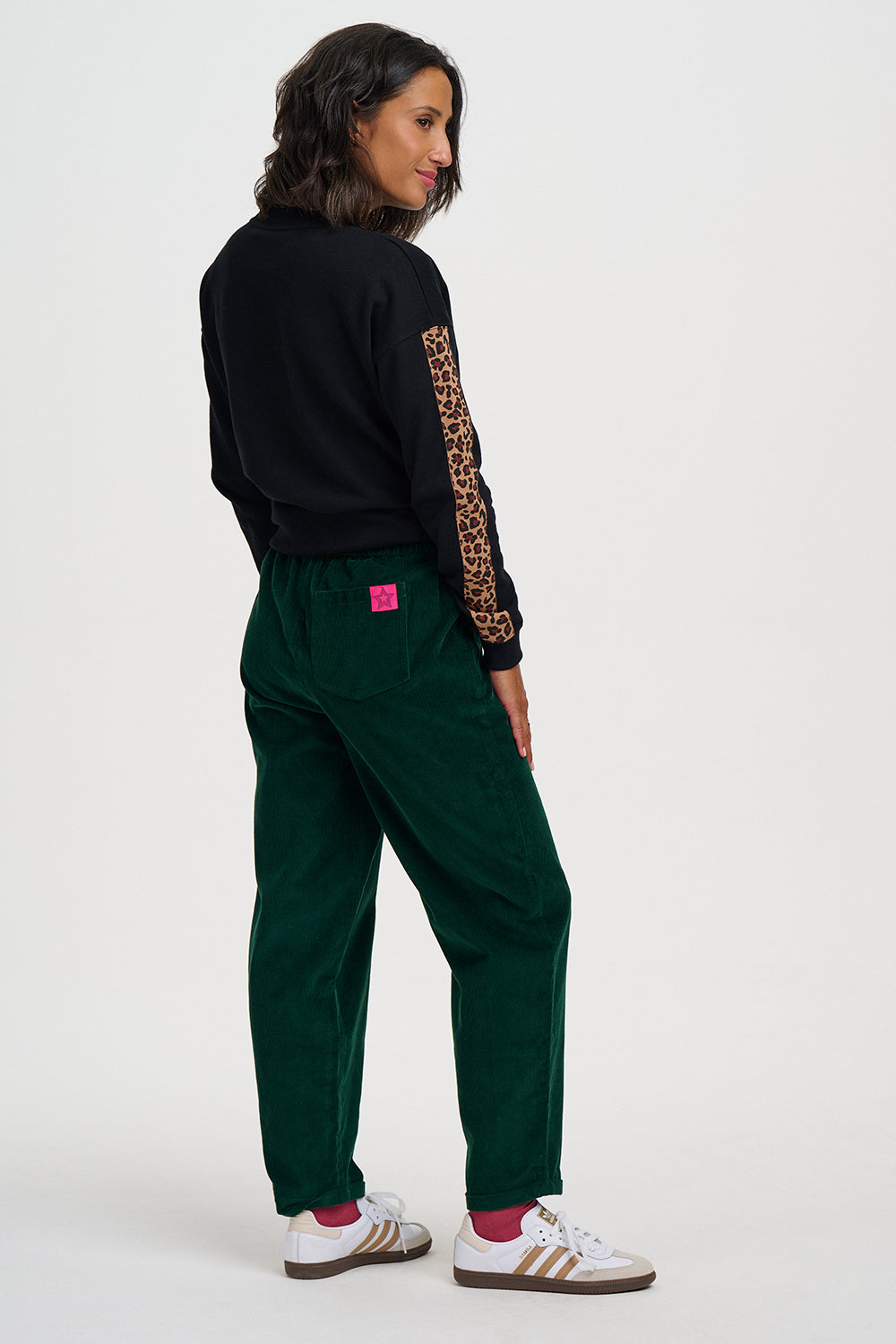 A woman shown from the back wearing dark green corduroy pants, a black sweatshirt with leopard-print sleeves, and white sneakers with pink socks, against a plain light gray studio background.