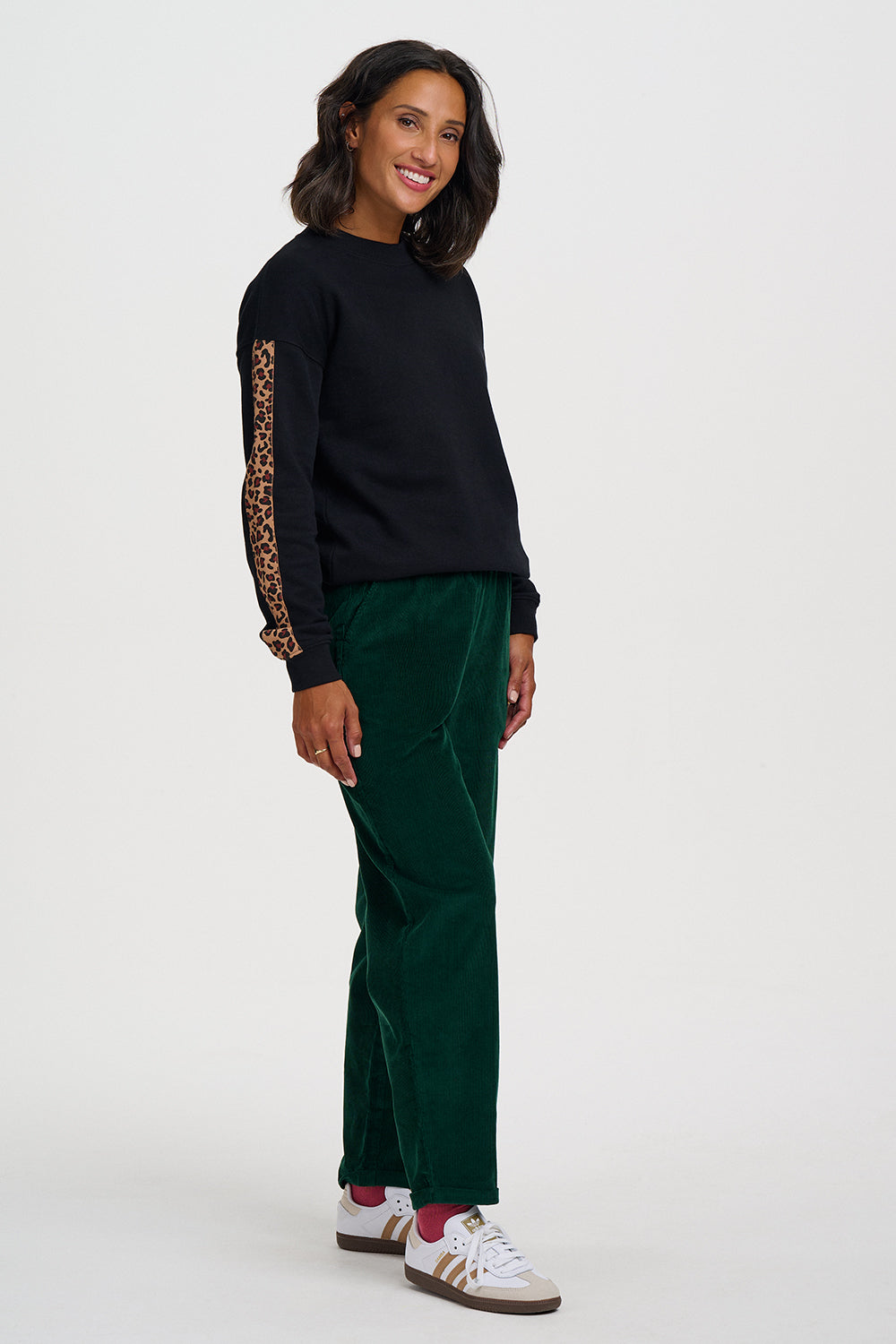 A woman modeling a black long-sleeve top with leopard-print sleeve panels and dark green trousers, standing against a light gray studio background.