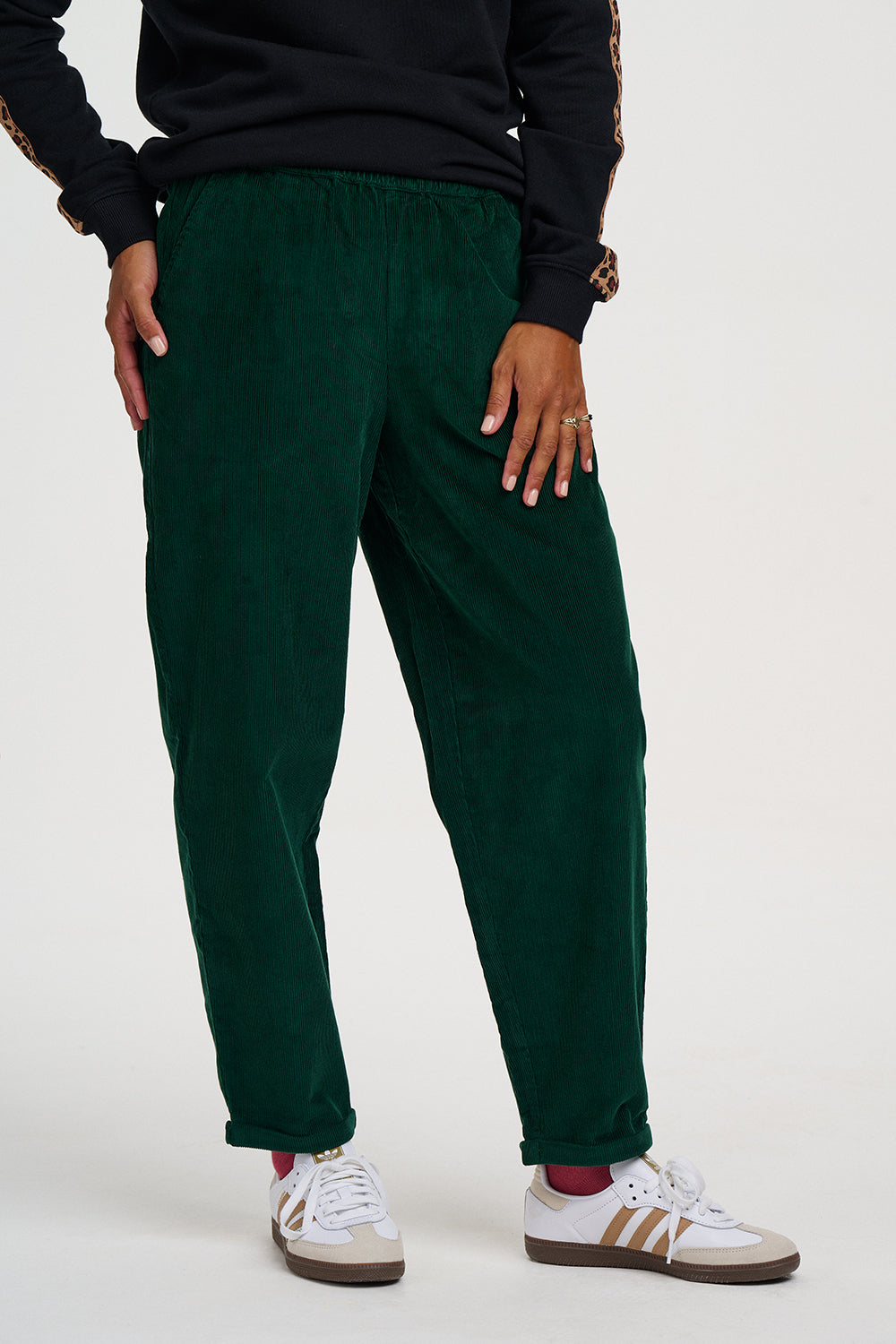 A person wearing dark green corduroy jogger pants with an elastic waistband, styled with a black sweatshirt and white sneakers with gold stripes, shown from the waist down on a light gray studio background.