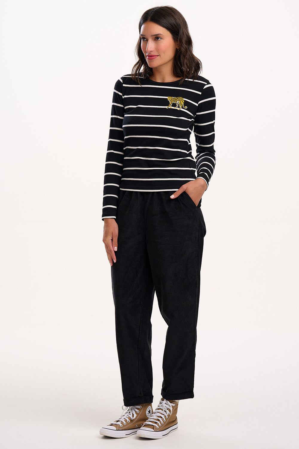 Woman wearing a black and white striped sweater with a yellow logo, black trousers, and brown sneakers on a white background.
