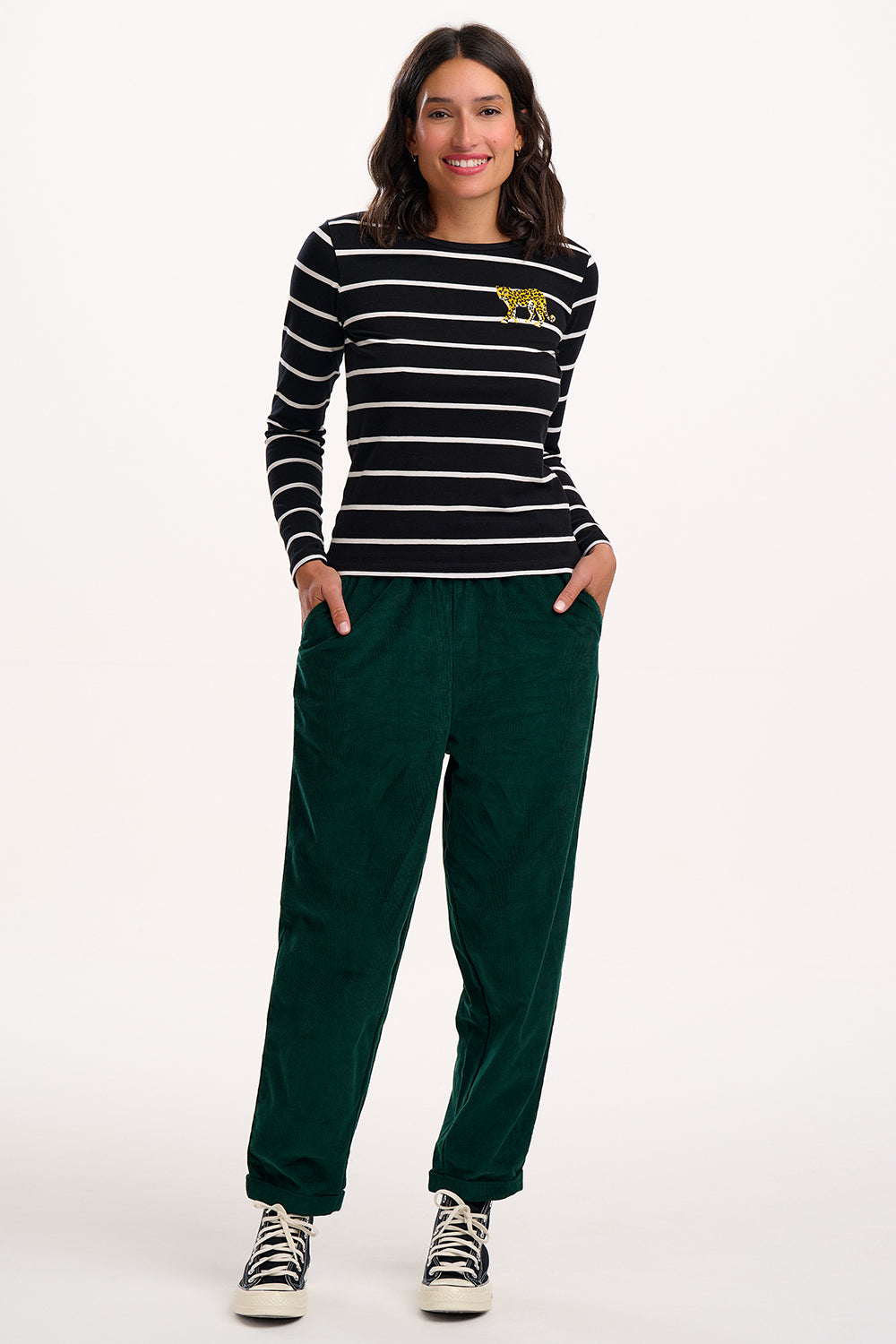 Woman wearing a black and white striped sweater with a yellow logo and green trousers on a white background